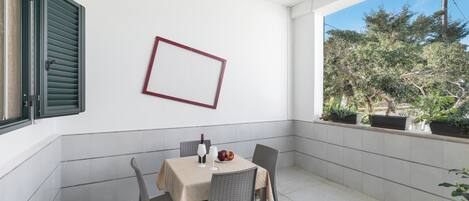 A dining area with a table set for two, a wicker chair, a hanging light fixture, and a view of trees through a window.