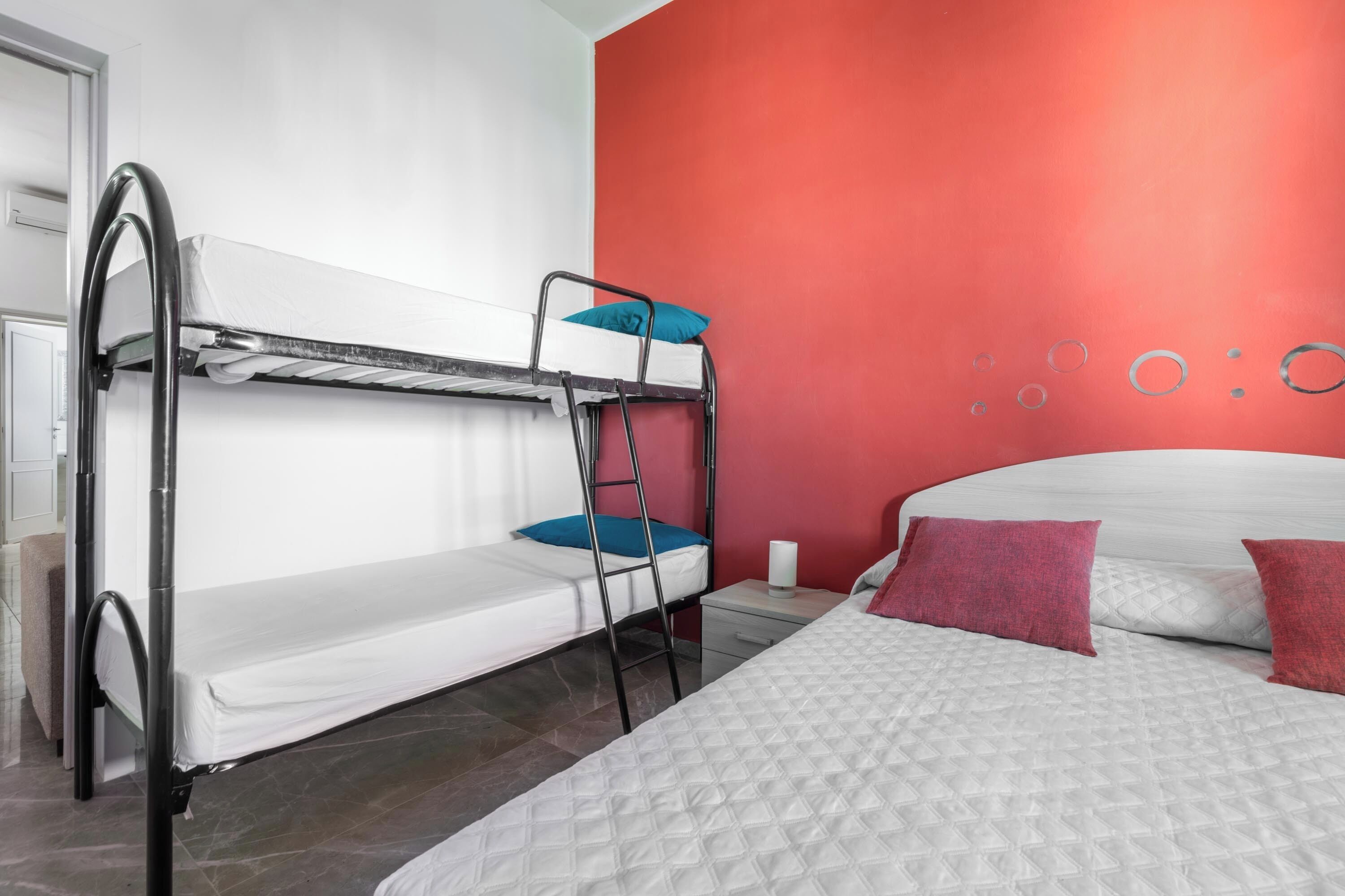 Habitación con literas, una con colchón blanco y la inferior con almohada azul. Hay una cama blanca con cubrecama acolchado y dos almohadas rojas. La habitación tiene una pared de acento roja y una mesita de noche blanca con una lámpara.