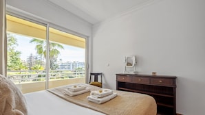 A hotel room with a large bed, a wooden dresser, and a view of palm trees and buildings through a large window. - Lux Garden View by Madeira Sun Travel (Funchal)