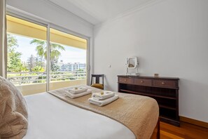 Une chambre d’hôtel avec un grand lit, une commode en bois et une vue sur les palmiers et les immeubles, visible à travers une grande fenêtre.