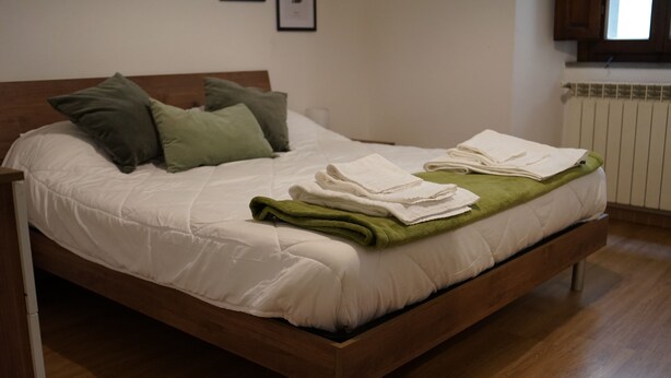 Un lit bien fait, avec une literie blanche et des coussins verts, un plaid vert et des serviettes blanches pliées, posés sur un cadre de lit en bois, dans une pièce au plancher en bois et dotée d’un radiateur.