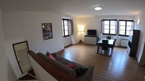 Un grand espace de vie comprenant un canapé, une table à manger et un téléviseur. De grandes fenêtres laissent entrer la lumière naturelle et offrent une vue sur l’extérieur.