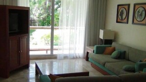 A living room with a sofa, a television, and a table. There is a window with curtains and a view of greenery outside. - Mayan Palace Vidanta Resort Riviera Maya 1 BR studio (Cancun)
