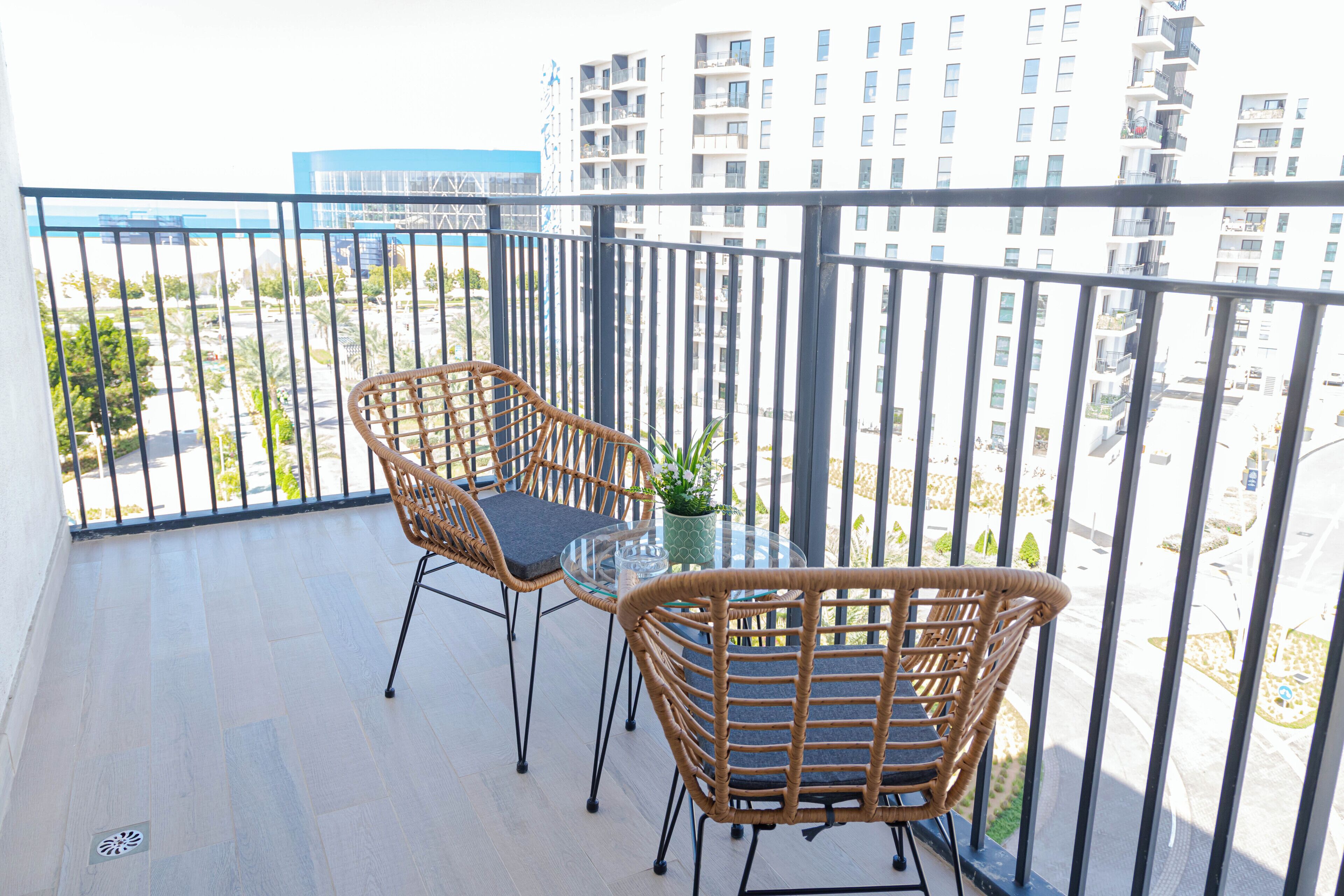 A balcony with wicker chairs and a glass table, overlooking a cityscape with high-rise buildings.