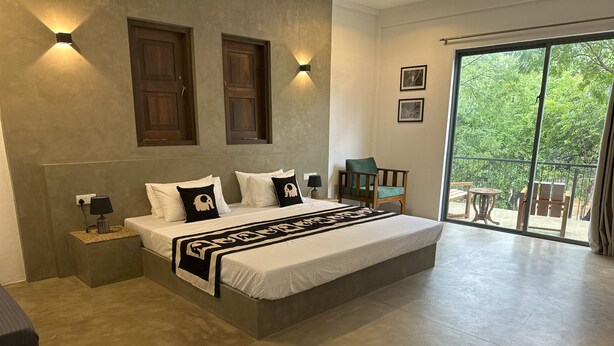 A large bed with a white and black bedspread, two nightstands with lamps, a wooden chair, and a view of greenery through a sliding glass door. - The Den at Yala  (Kataragama)