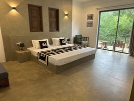 Una cama grande con colcha blanca y negra, dos mesitas de noche con lámparas, una silla de madera y vistas a zonas verdes a través de un ventanal corredero.
