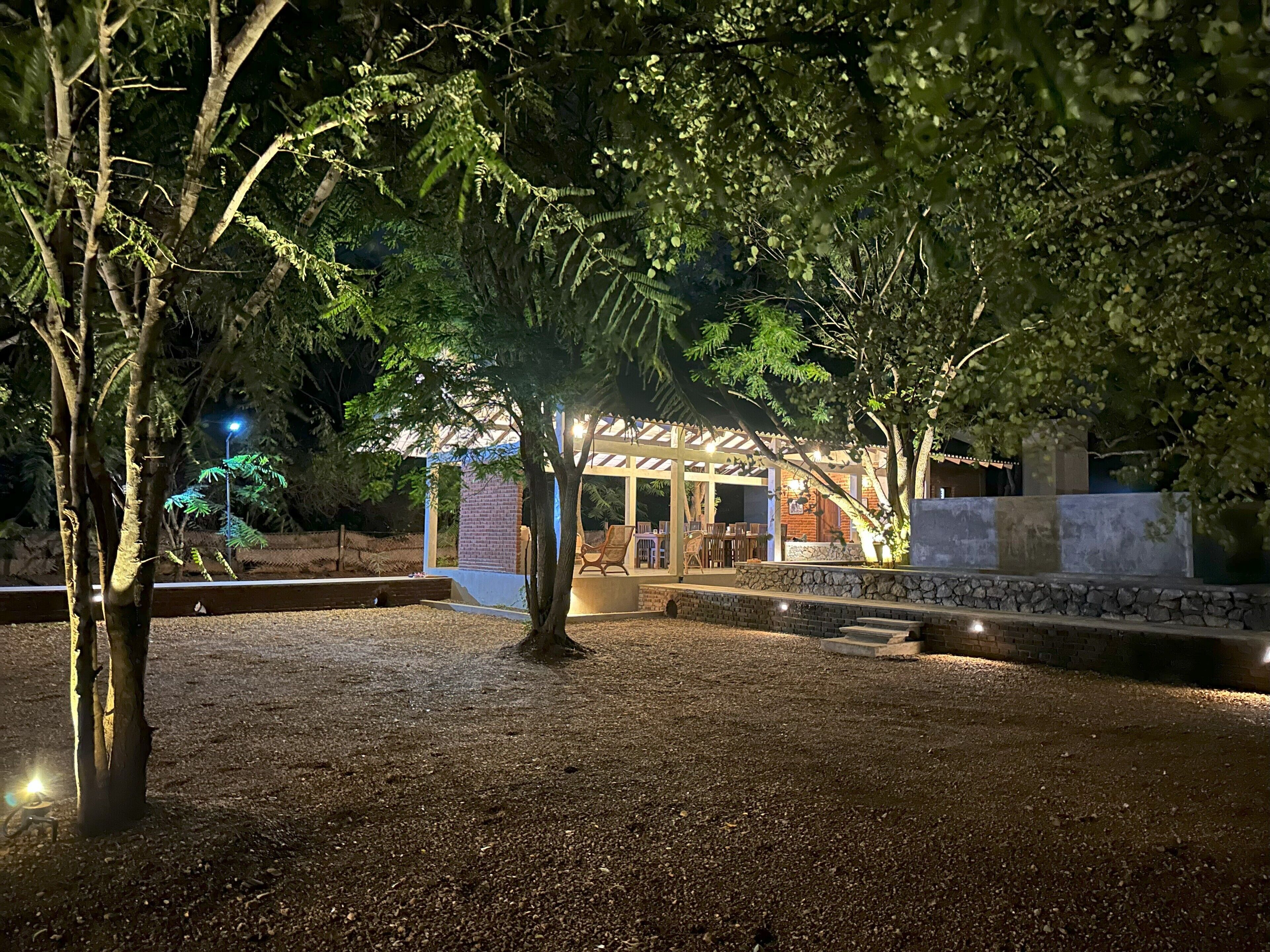 Un espacio de estar al aire libre cubierto, con paredes de piedra y una estructura de pérgola, rodeado de árboles y grava.