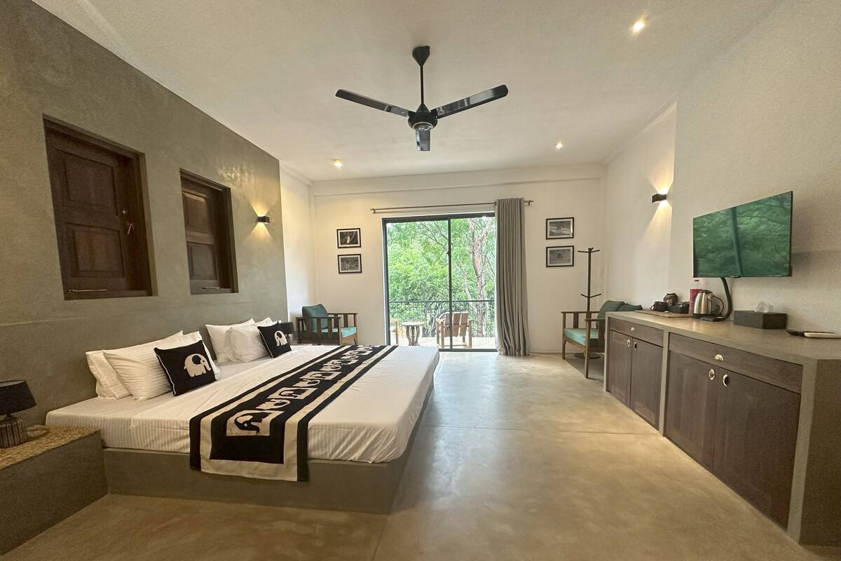 A modern hotel room with a large bed, a desk, and a balcony with a view of trees.