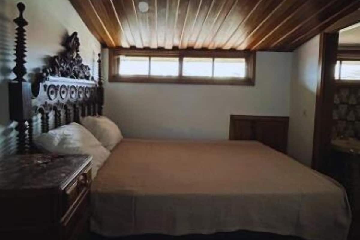 A bedroom with a wooden ceiling, a large bed, a nightstand, and a window with blinds.