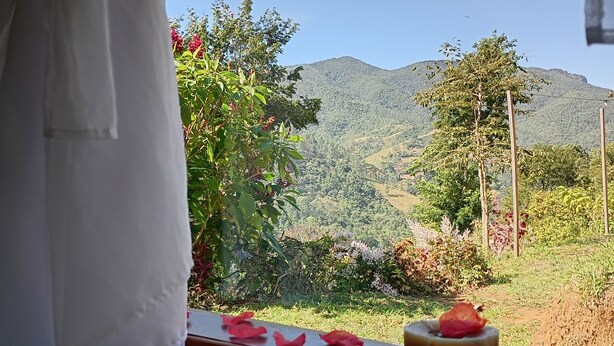 Una finestra con vista su un paesaggio montuoso, una panca di legno con un cuscino e un tavolino con una candela e petali di rosa.