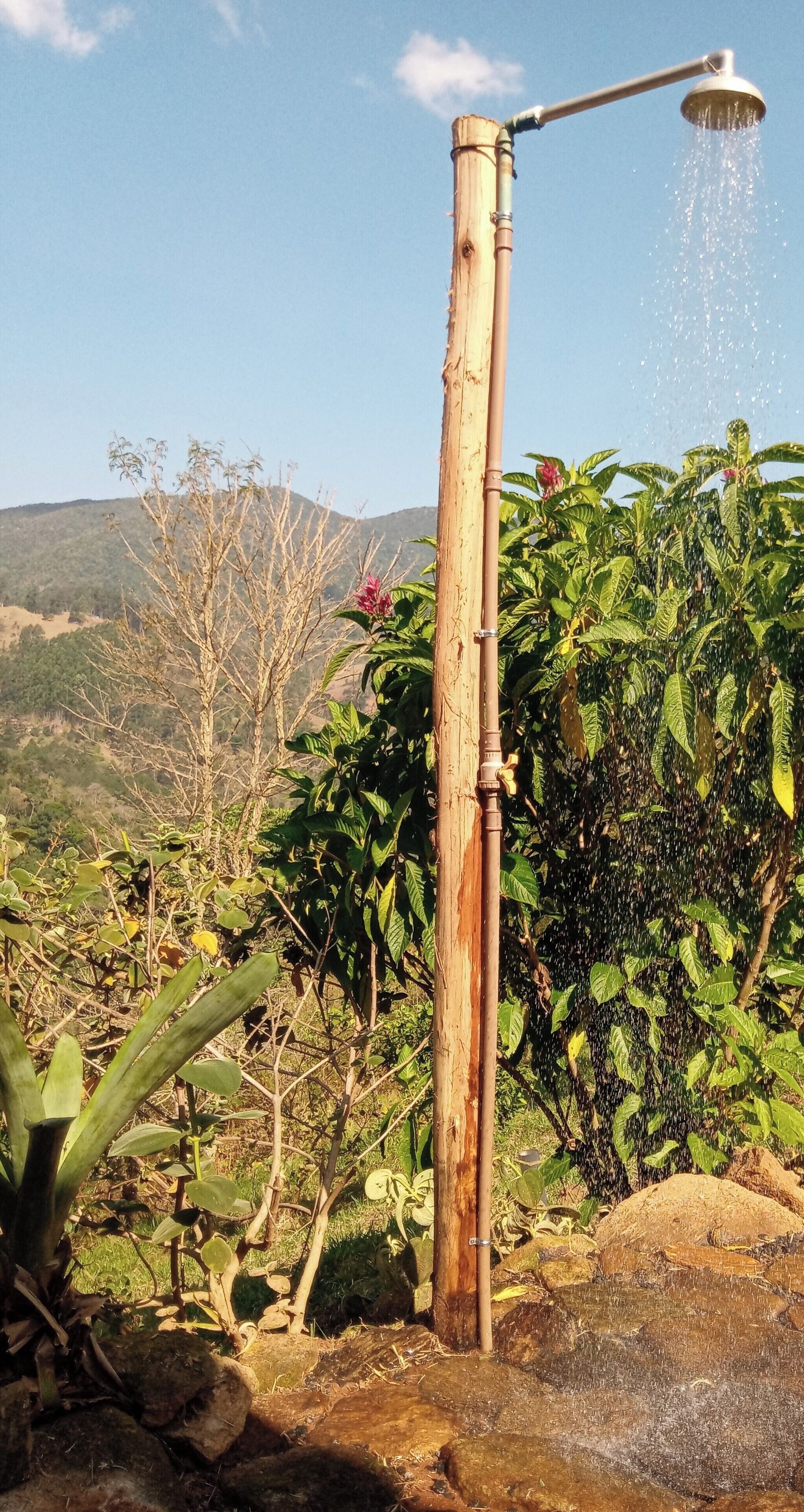 A bamboo showerhead with water spraying down, set against a backdrop of lush greenery and a clear sky.