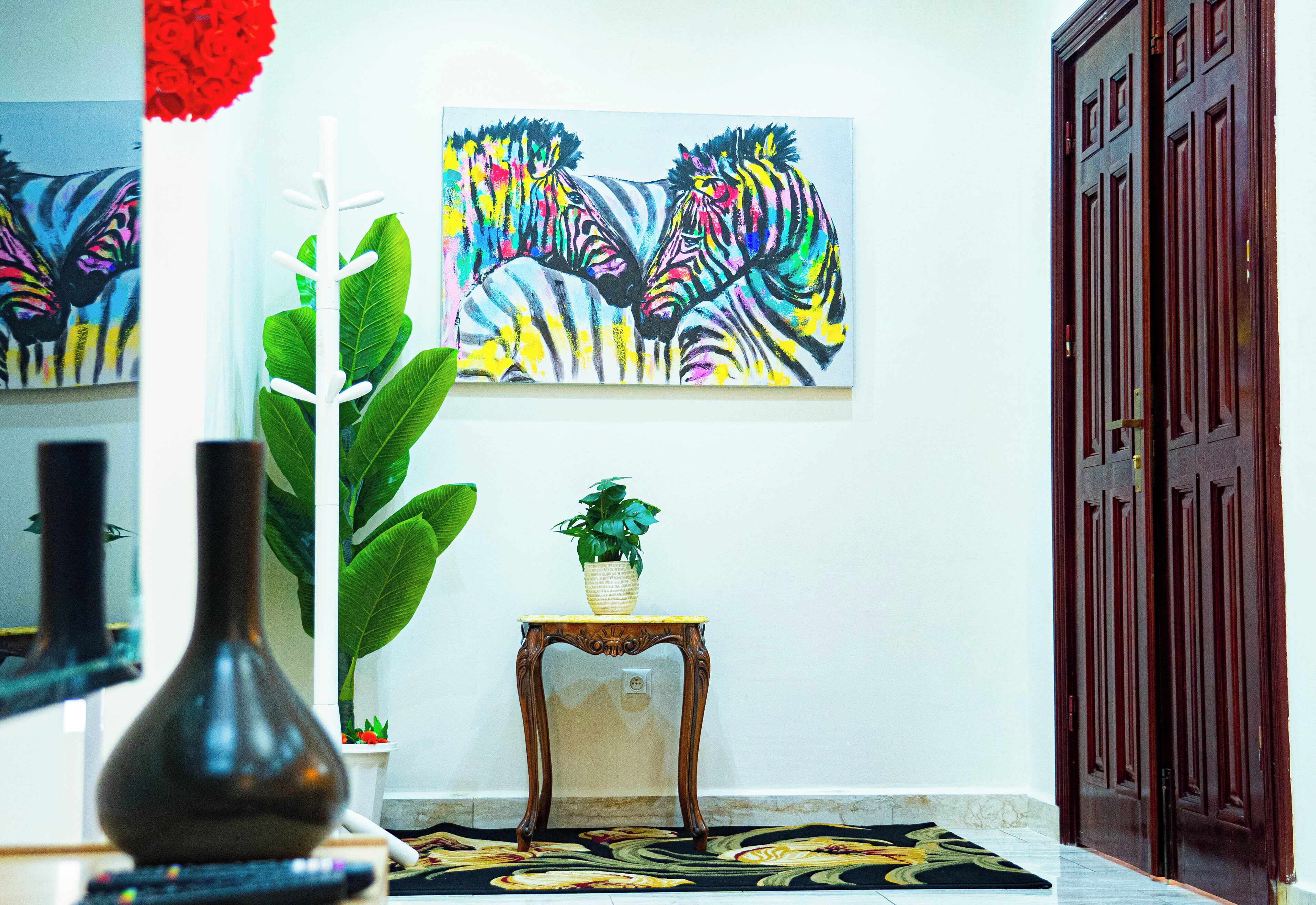 A room with a large painting of zebras, a potted plant, a vase, and a wooden table.
