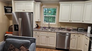 A kitchen with white cabinetry, a stainless steel refrigerator, a dishwasher, a granite countertop, and a window with a view of trees.
