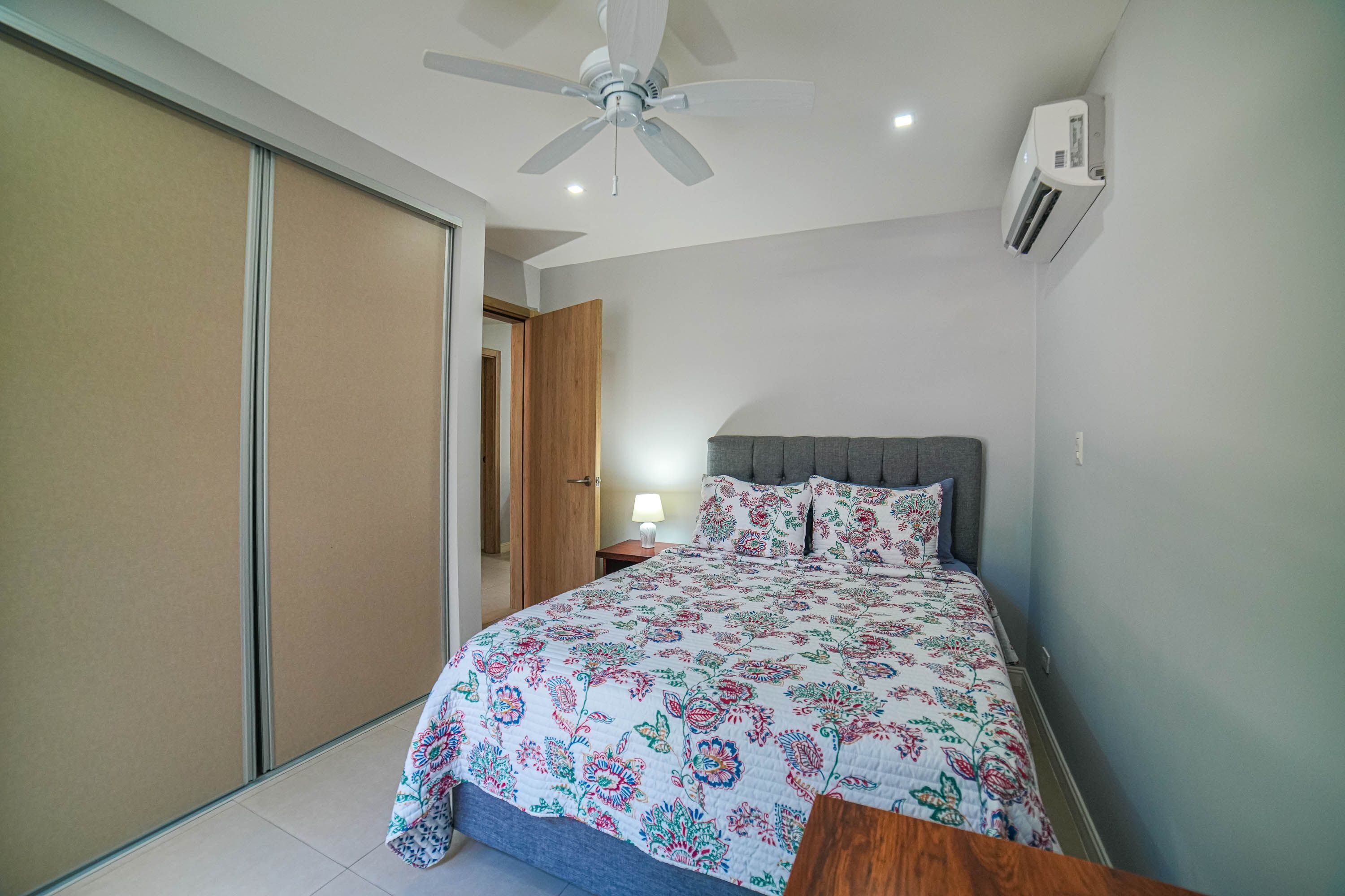 A bedroom with a bed, a ceiling fan, an air conditioning unit, and a wooden nightstand.