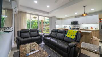 Una sala de estar moderna con un sofá de cuero, una mesa de centro de vidrio y una zona de cocina con armarios blancos y un microondas.