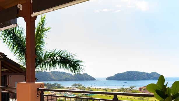 Un balcon avec vue sur une plage, des palmiers et des collines lointaines.