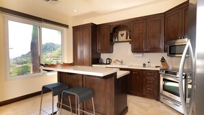 Une cuisine moderne avec des meubles de couleur bois foncé, un grand îlot central doté de tabourets, un four encastré et une vue sur la verdure à l’extérieur.