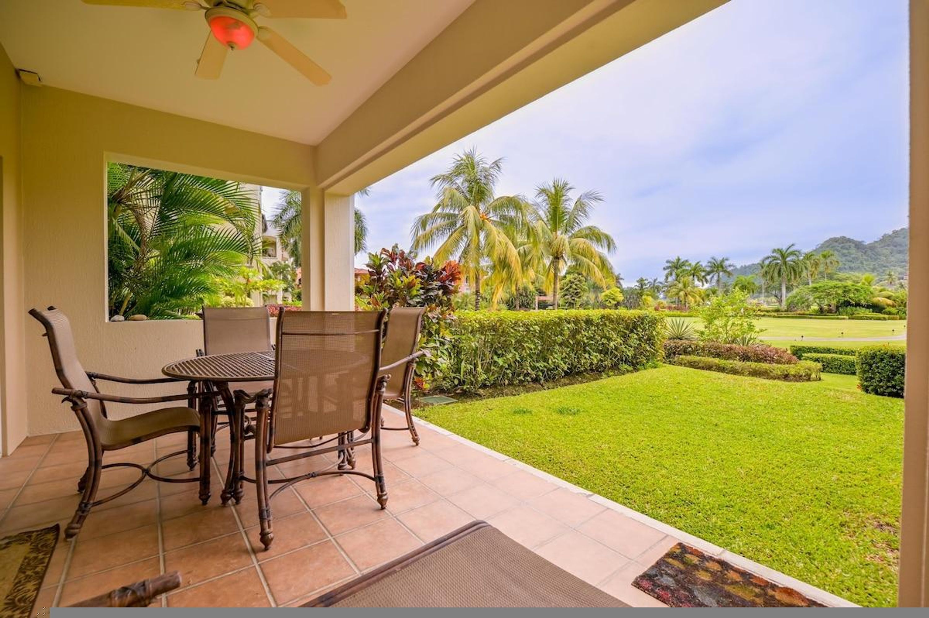 Un patio techado con una mesa y sillas, vistas a un césped cuidado y palmeras, y un ventilador de techo.