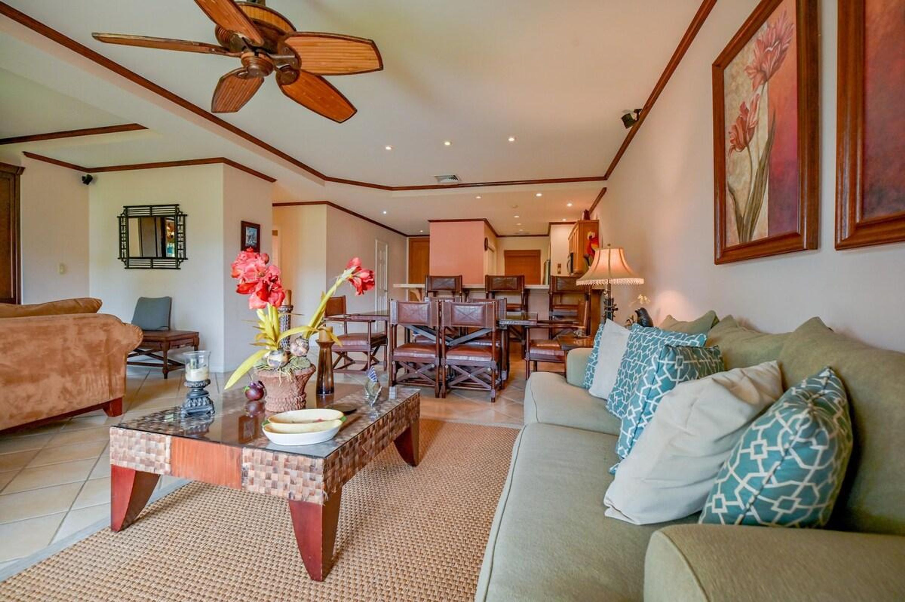 Ein geräumiges Wohnzimmer mit Deckenventilator, Couchtisch mit Dekoration, Sofa mit gemusterten Kissen und Essbereich mit Holztischen und Stühlen.