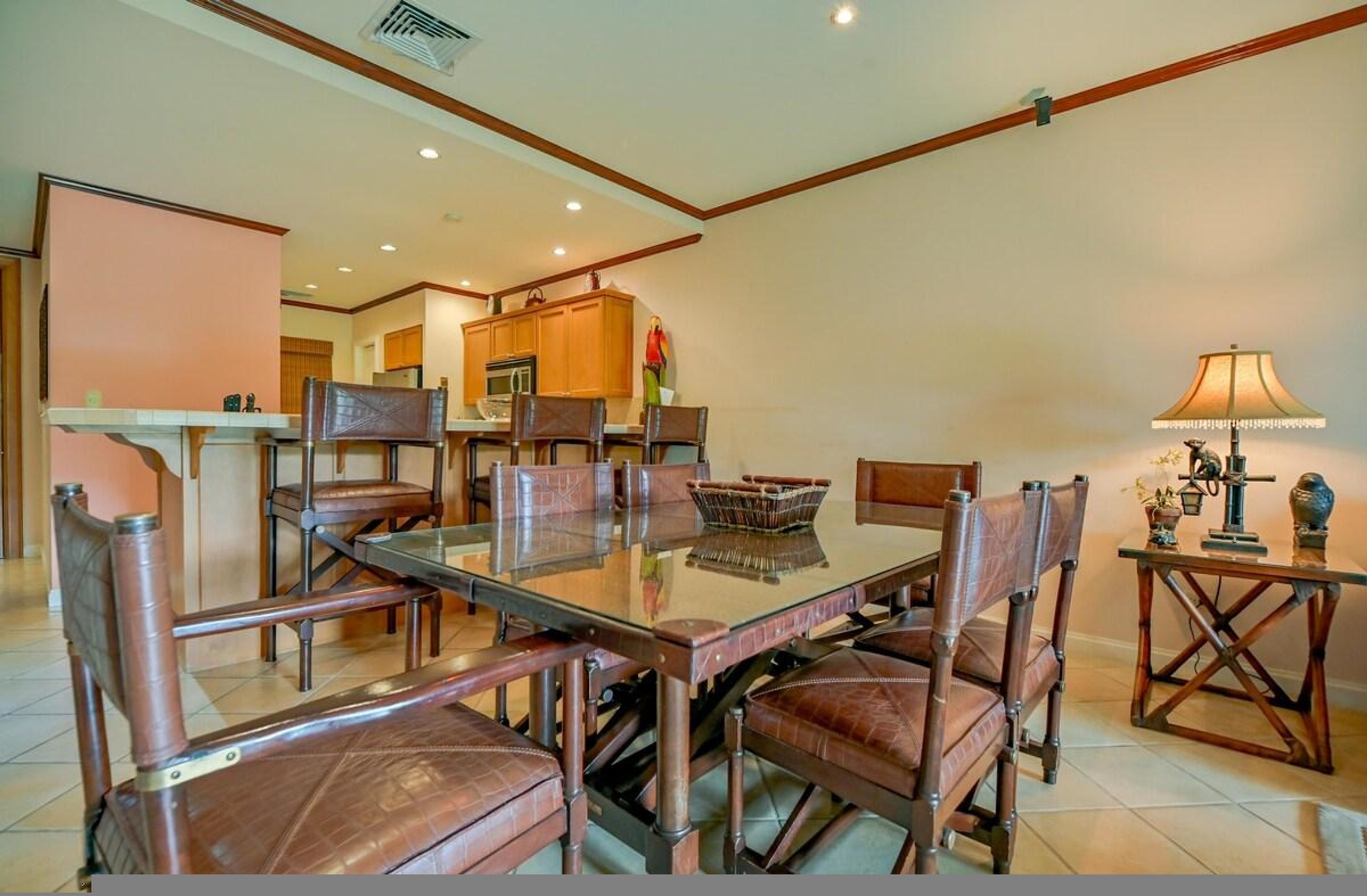 A dining area with a glass-top table surrounded by leather chairs, wooden cabinets, and a kitchen in the background.