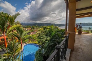 A balcony with a view of a swimming pool, lush greenery, and distant mountains.