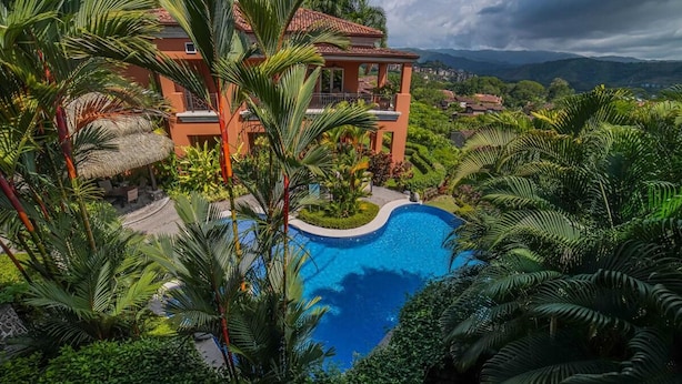 Un complexe hôtelier doté d’une piscine entourée d’une végétation luxuriante et d’une grande maison avec un toit rouge.