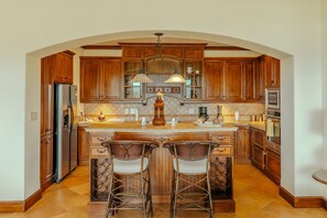 A kitchen with wooden cabinets, a stainless steel refrigerator, a large island with two bar stools, and a hanging light fixture. - CR Luxury Montebello 1D 3 Bdrm (Jacó)