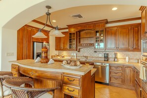 A spacious kitchen with wooden cabinets, a large island with a sink, and a dining area with wicker chairs. - CR Luxury Montebello 1D 3 Bdrm (Jacó)