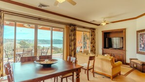 Amplia sala de estar con mesa de comedor, sofás y televisor. Ventanas grandes ofrecen vistas al exterior.