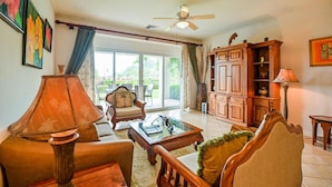 Uma sala de estar ampla com sofá, poltrona e mesa de centro. Há um armário de madeira e um ventilador de teto.