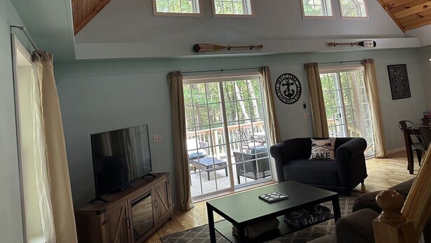 A spacious living room with a high ceiling, large windows, a television, a sofa, and a dining area visible through the glass doors. - Great Lake Haven between Pentwater and Silver Lake (Mears)