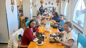 A group of people dining in a restaurant with a long table, various dishes, and a view through large windows. - New Abusimble Hotel (Abu Simbel)