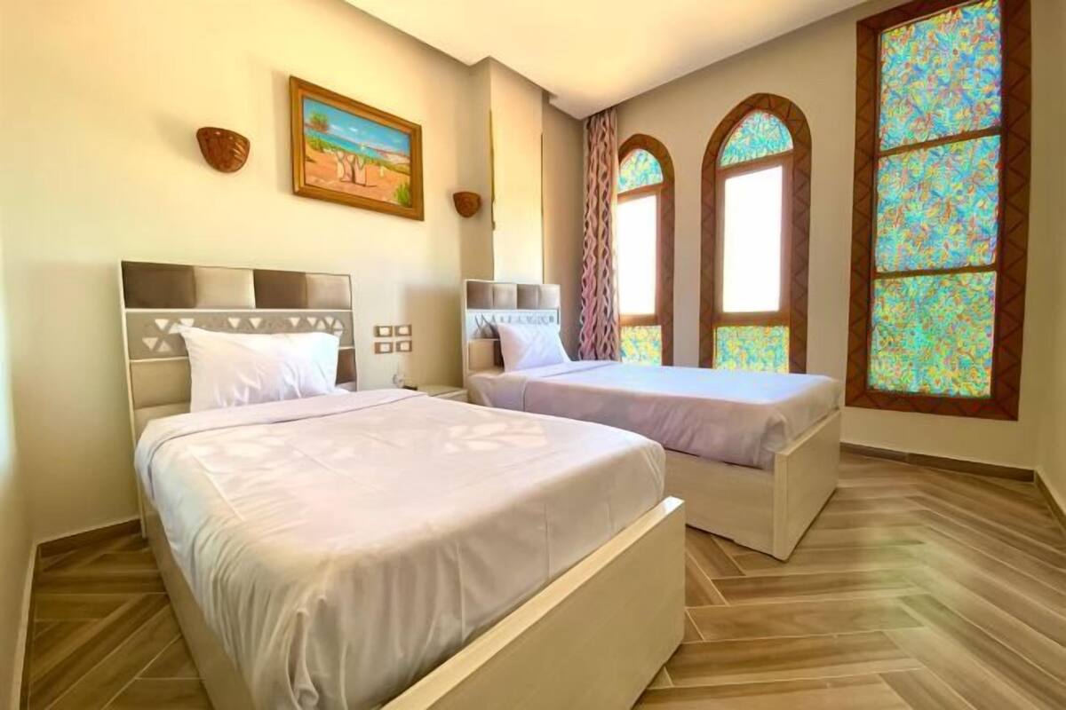 A hotel room with two beds, a painting on the wall, and stained glass windows.
