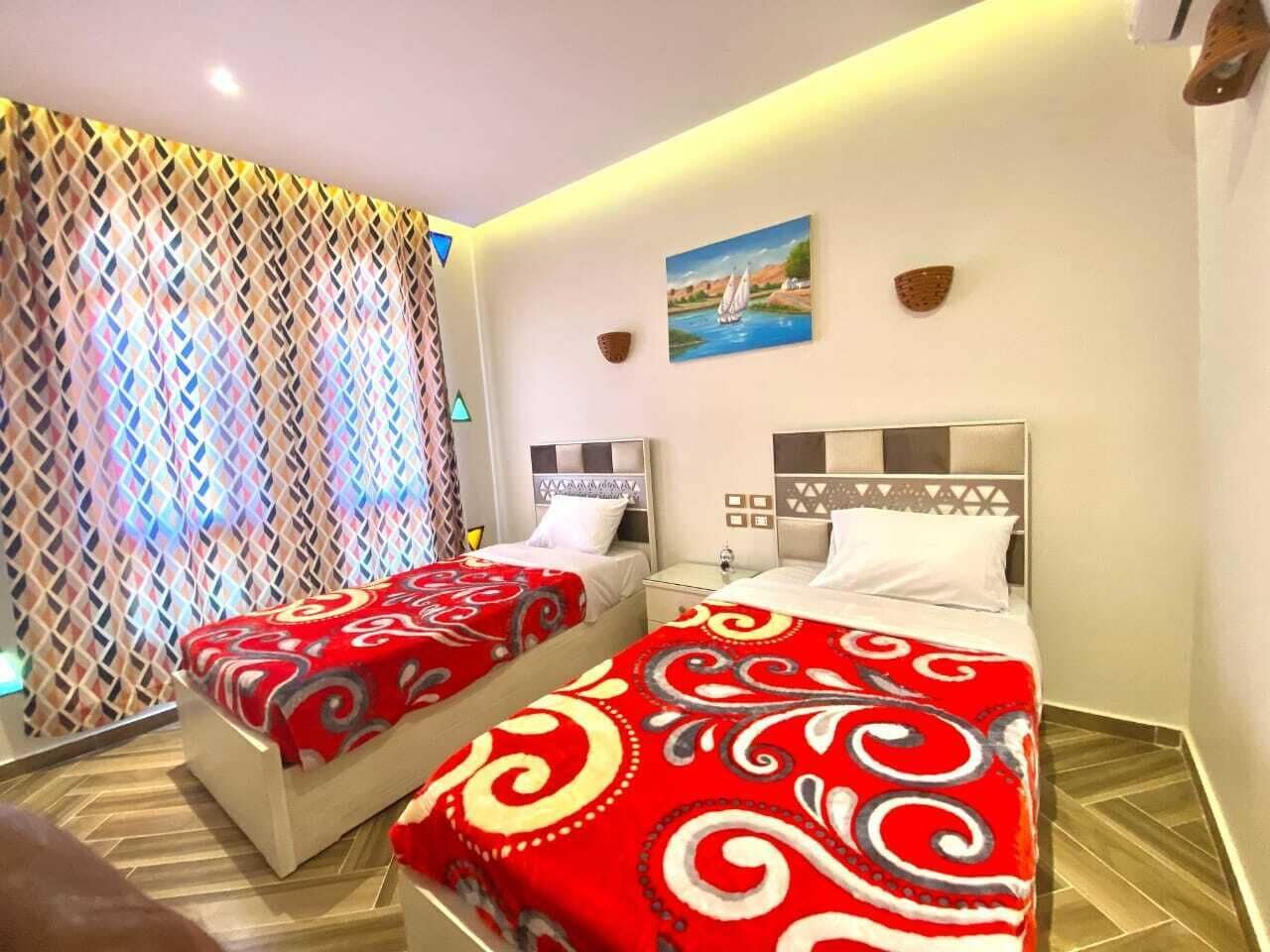 Ein Hotelzimmer mit zwei Betten, jeweils mit einer rot-weißen Musterdecke. An der Wand hängt ein Gemälde, das eine Landschaft mit einem Boot zeigt.