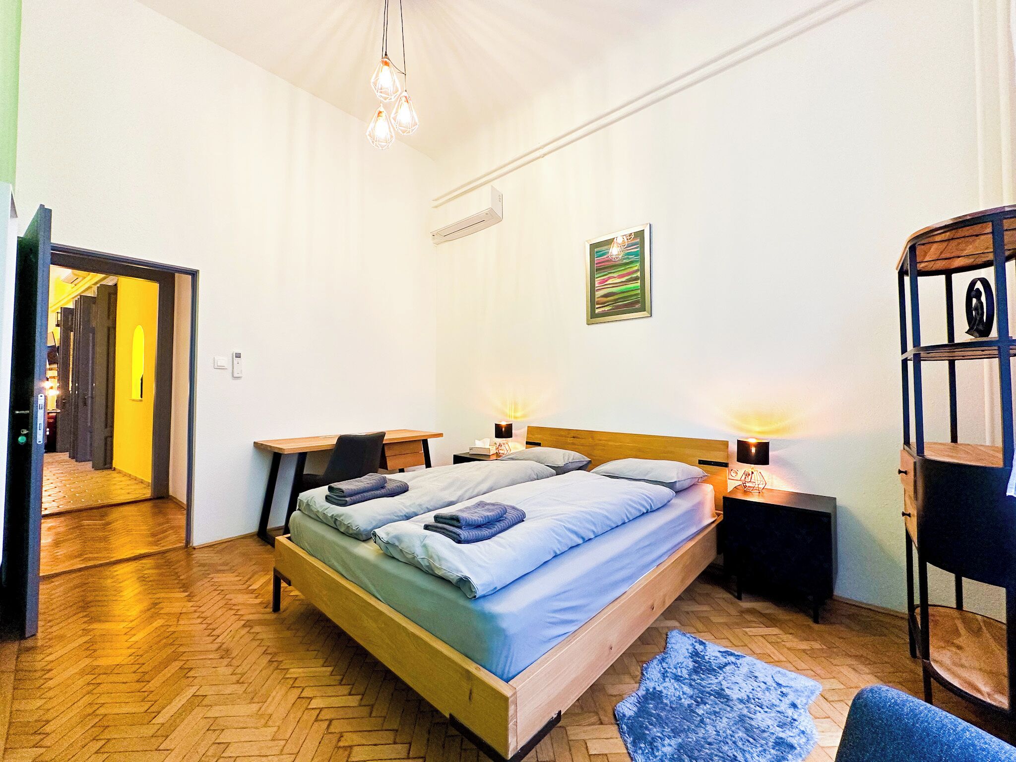 A bedroom with a bed, bedside tables, a desk, a chair, a hanging light fixture, and a wall-mounted air conditioner.