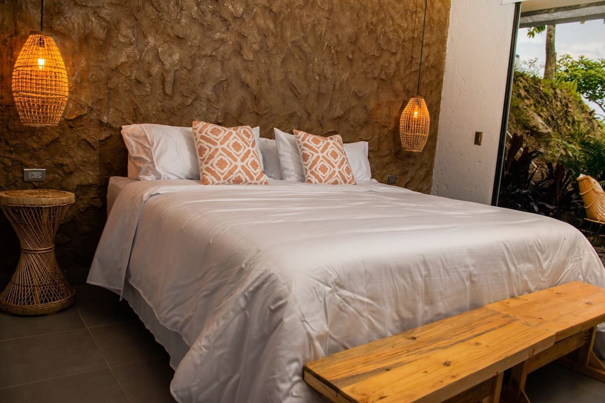 A bed with white bedding and patterned pillows, a wooden bench, wicker lamps, and a stone wall.