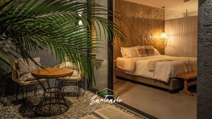 A bedroom with a bed, bedside table, and a small table with chairs. There is a large plant and a ceiling fan. - Santuario Luxury Ecohotel (Villeta)
