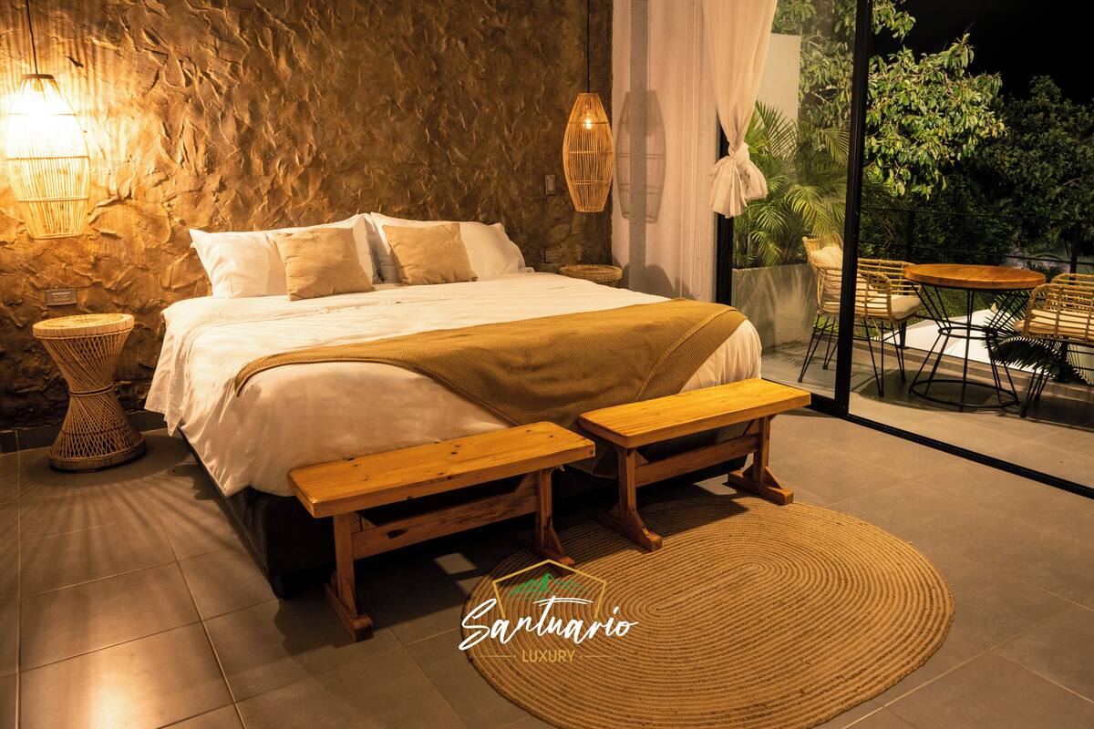 A bedroom with a large bed, bedside tables, a bench, and a view of the outdoors.