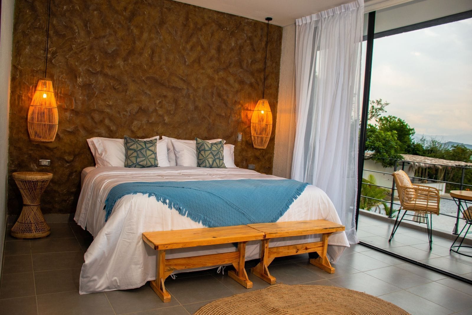A bedroom with a large bed, two bedside lamps, a wooden bench, and a view of the outdoors.