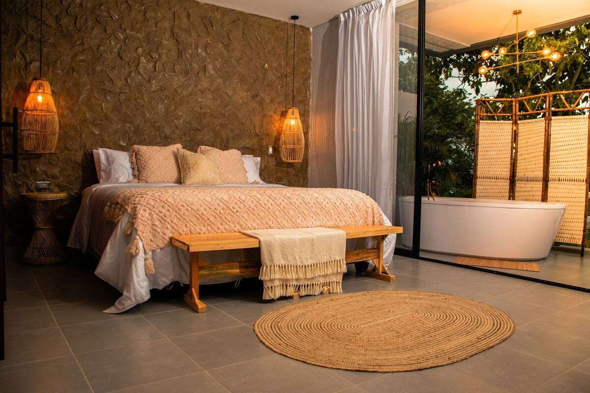 A modern bedroom with a stone wall, a large bed, bedside lamps, a bench, a round woven rug, and a bathtub.