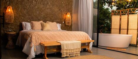A modern bedroom with a stone wall, a large bed, bedside lamps, a bench, a round woven rug, and a bathtub.