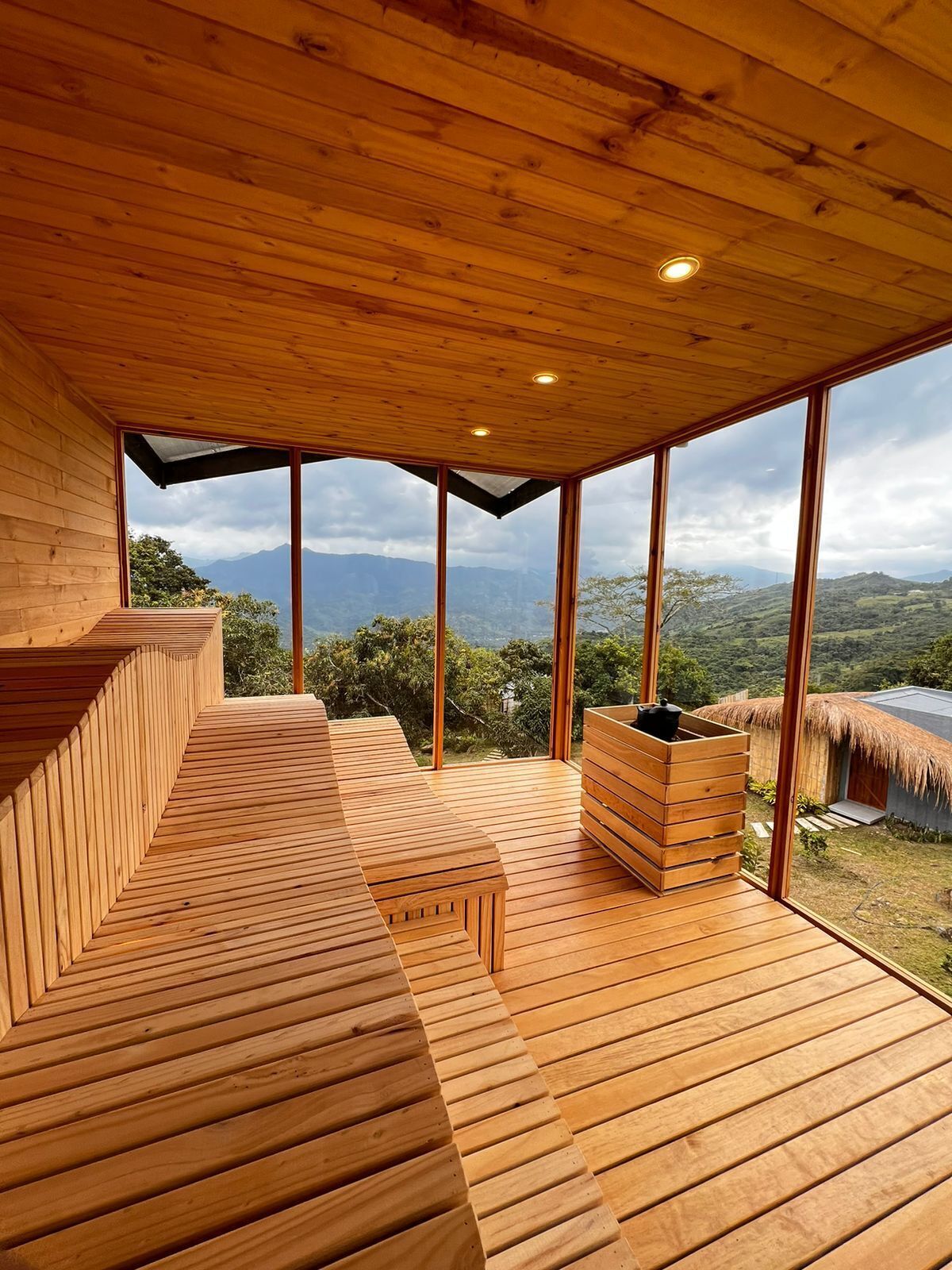 A wooden interior with large windows offering a view of mountains and a thatched-roof structure outside.