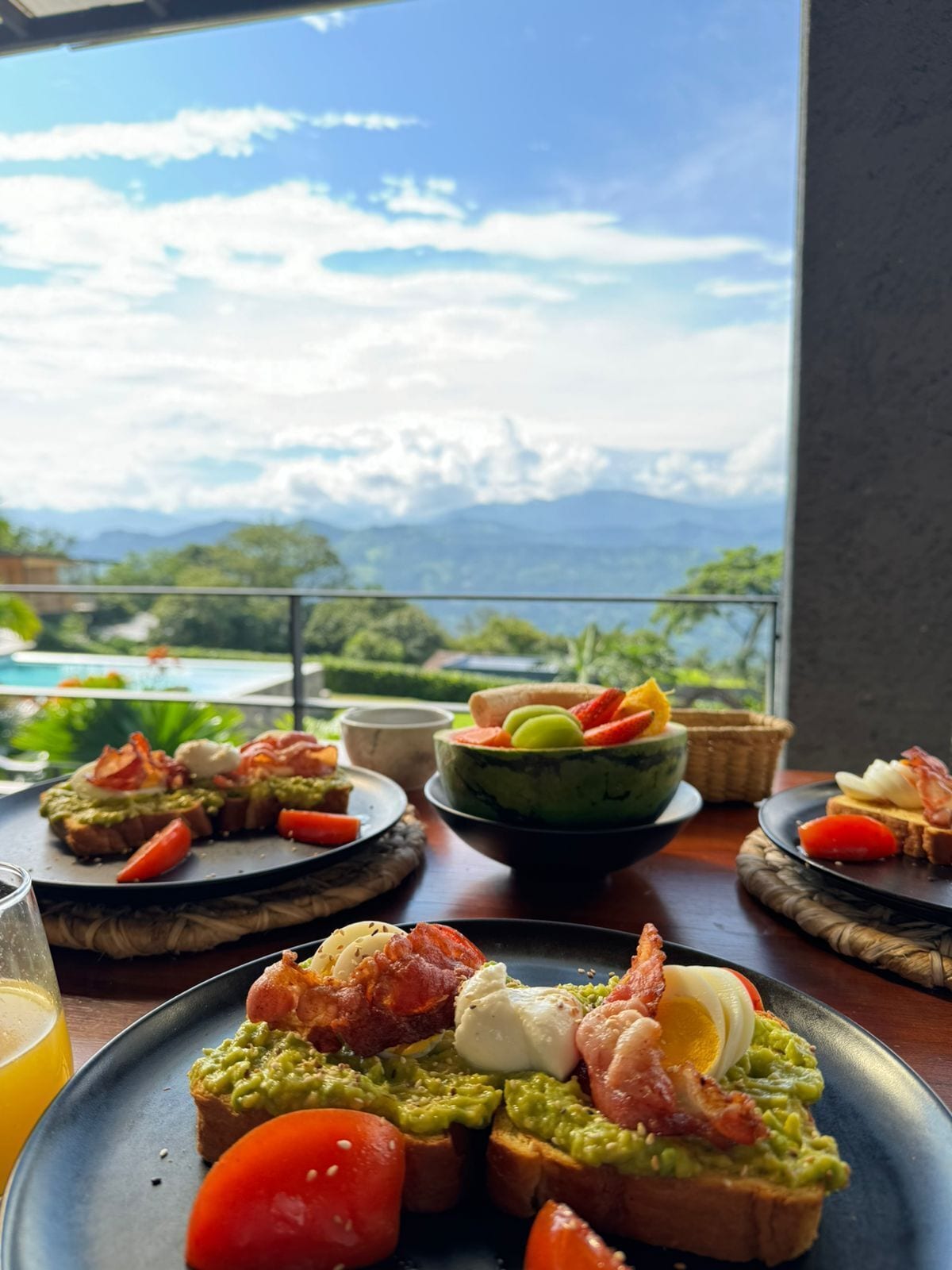 Una mesa con diversos alimentos para el desayuno, incluyendo tostadas con aguacate y tocino, un bol de fruta y un vaso de jugo de naranja.