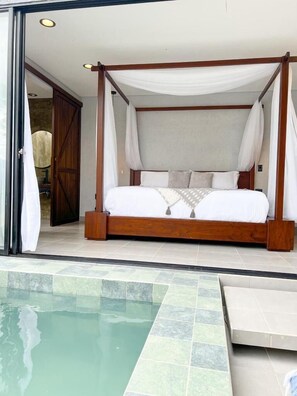 Una cama con dosel de madera y ropa blanca, ubicada junto a una piscina con piso de baldosas.