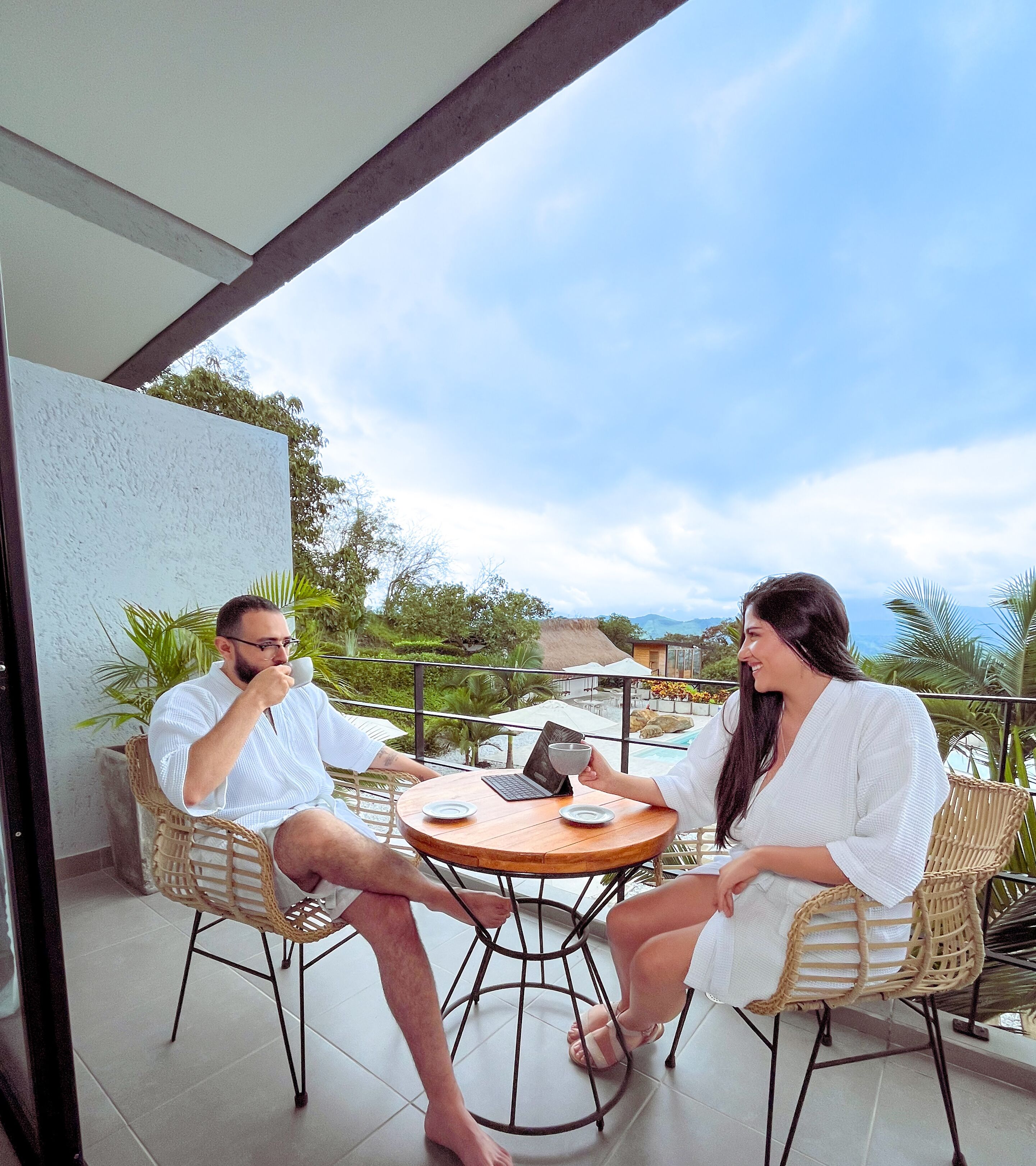 Due persone sedute a un tavolo rotondo su un balcone con vista su una piscina e vegetazione.