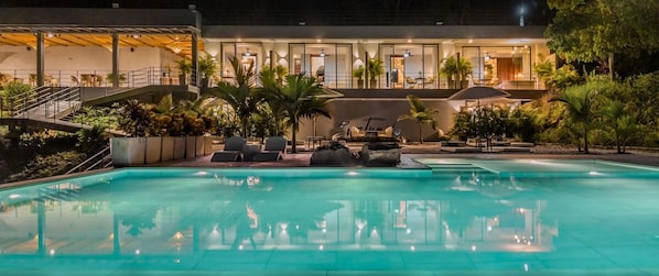 Una piscina con sillas de descanso y sombrillas, rodeada de vegetación y un edificio de varios pisos con amplias ventanas.