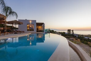 Una piscina a sfioro moderna con vista libera sul mare al tramonto, un’area relax con ombrelloni e un edificio contemporaneo con ampie vetrate.