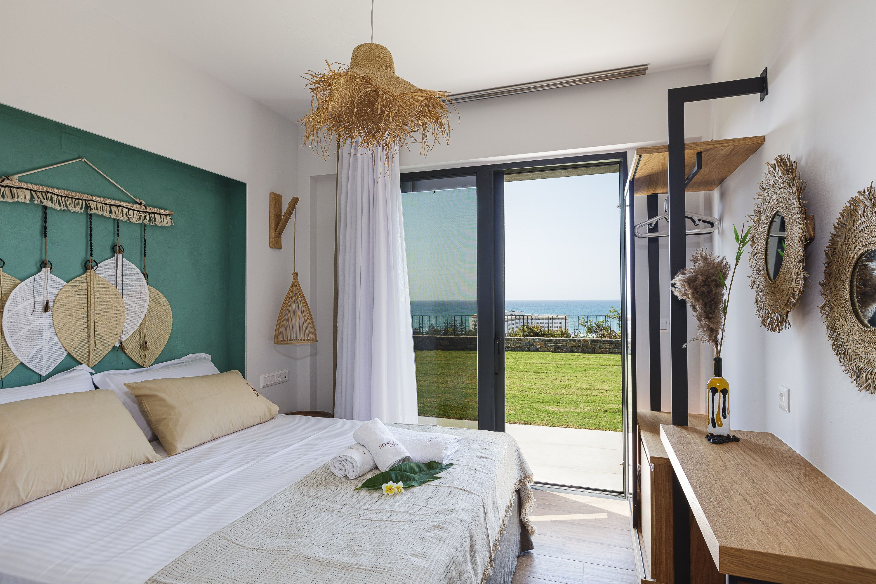 A modern bedroom with a large bed, a view of the sea, and a balcony with a wooden table and decorative mirrors.
