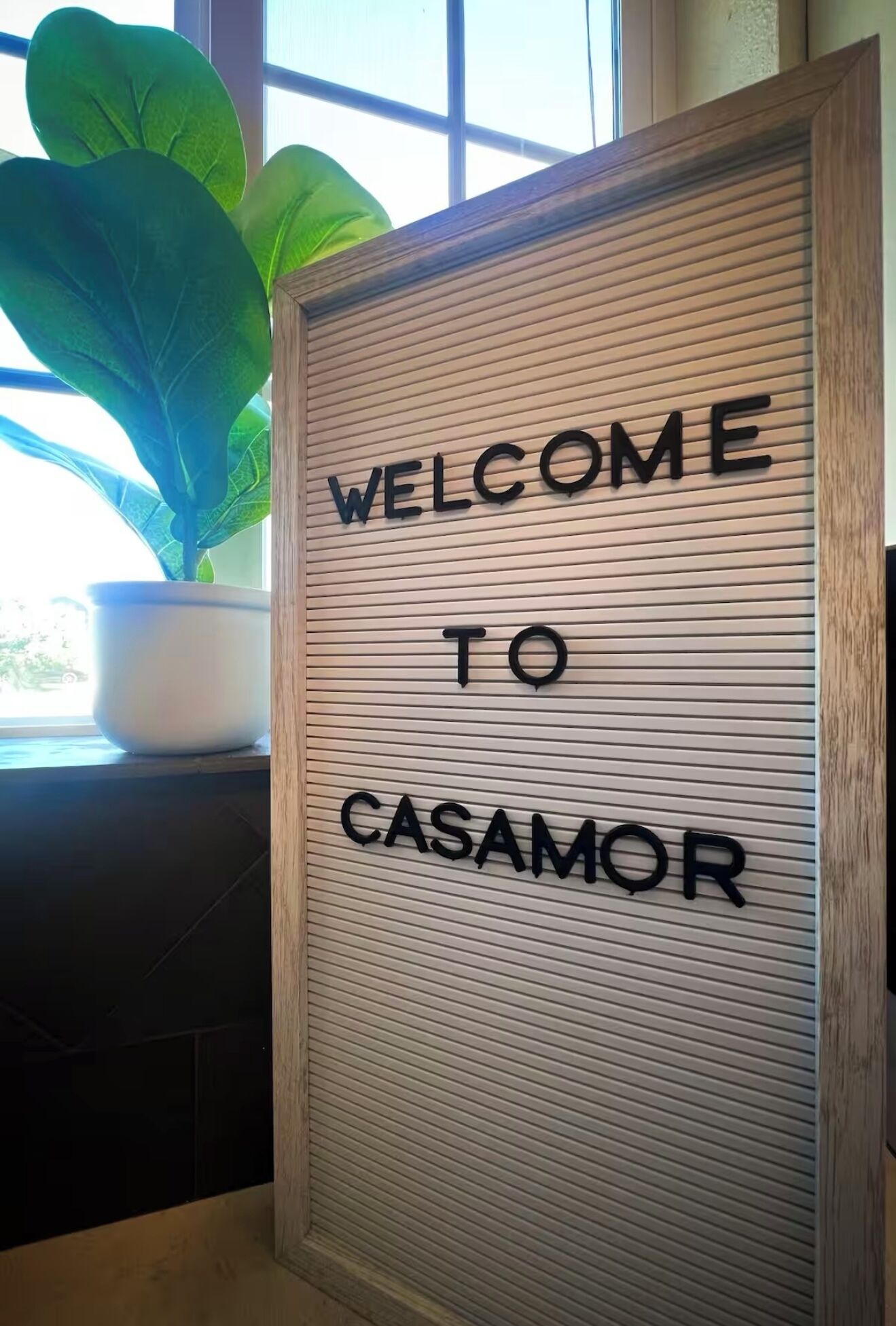 A framed welcome sign with the text "WELCOME TO CASAMOR" against a window with blinds.