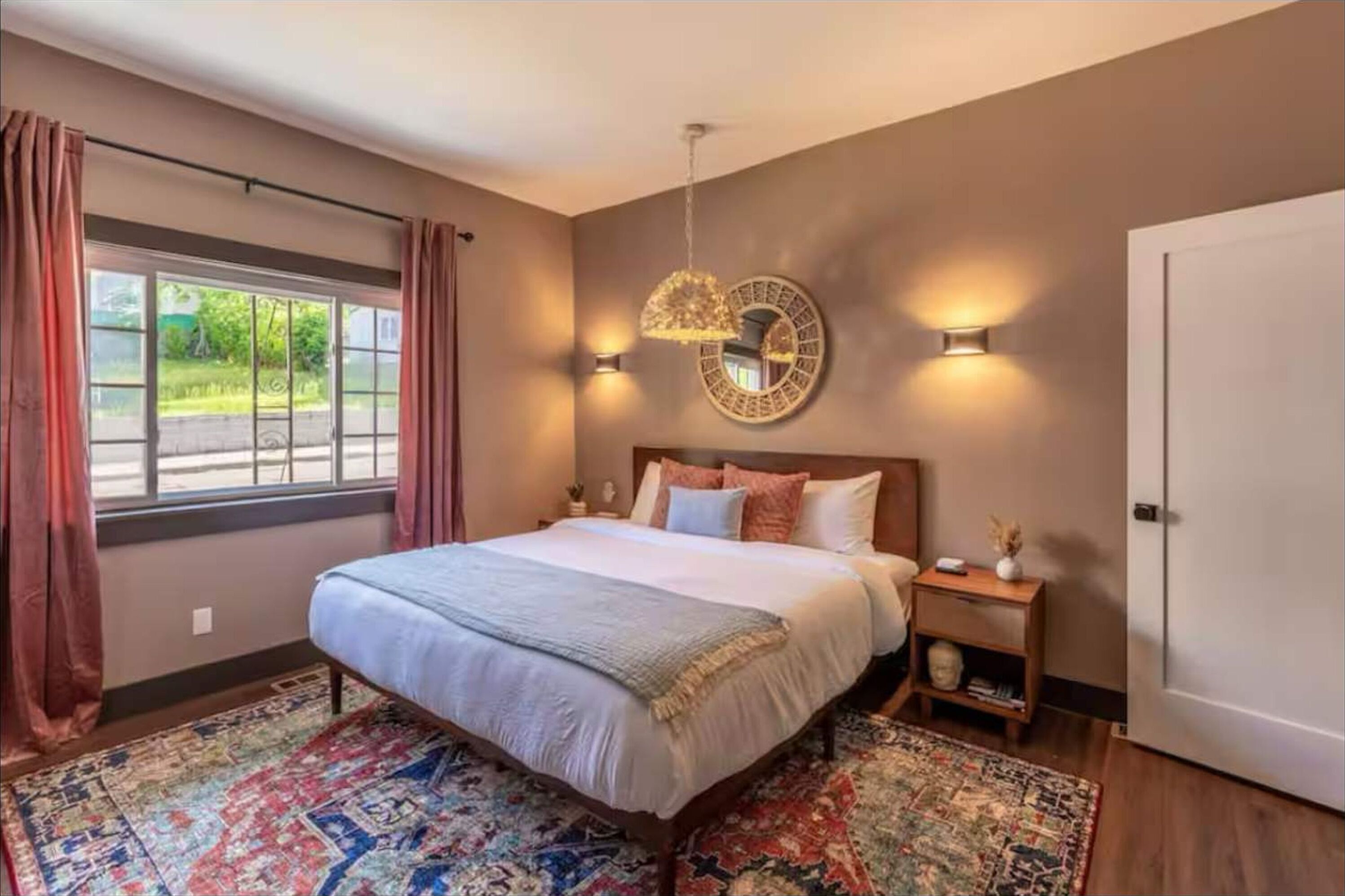 Une chambre avec un grand lit, un tapis à motifs, une table de chevet avec une lampe et une vue sur l’extérieur par une fenêtre.